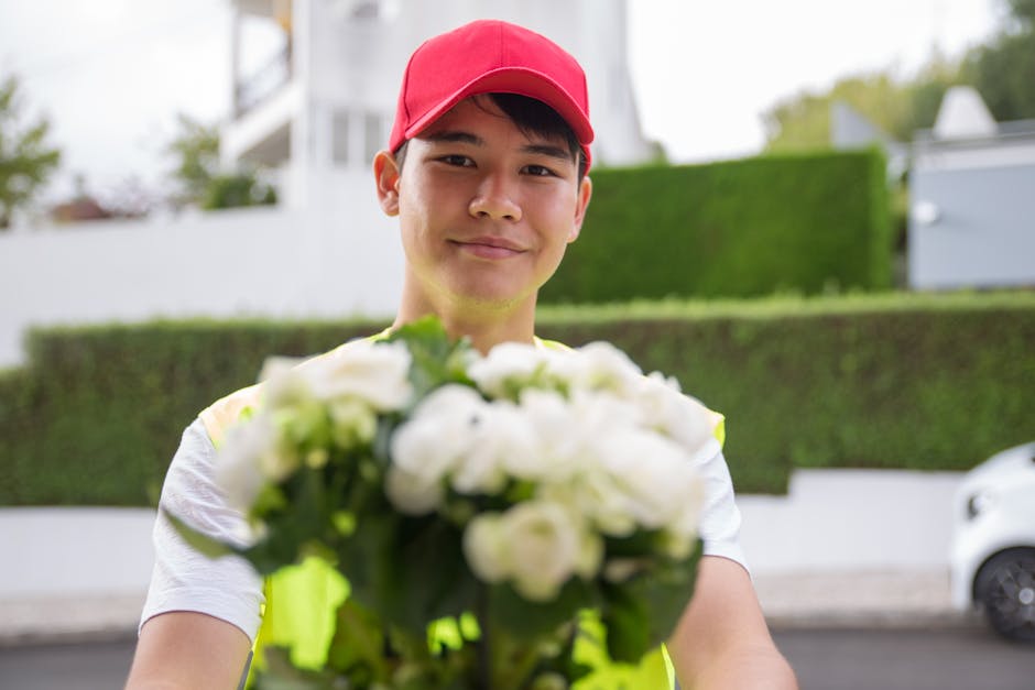 A young male flower delivery courier with light skin, wearing a bright red cap and a high-visibility vest, holding a bouquet of fresh white flowers with green leaves towards the camera. The bouquet ap