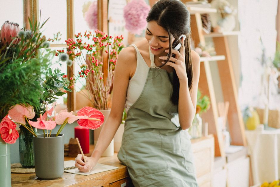 A young woman smiling while talking on a mobile phone inside a floral shop, standing behind a wooden counter. She is wearing a light grey apron over a white sleeveless top and has long, dark hair. In 