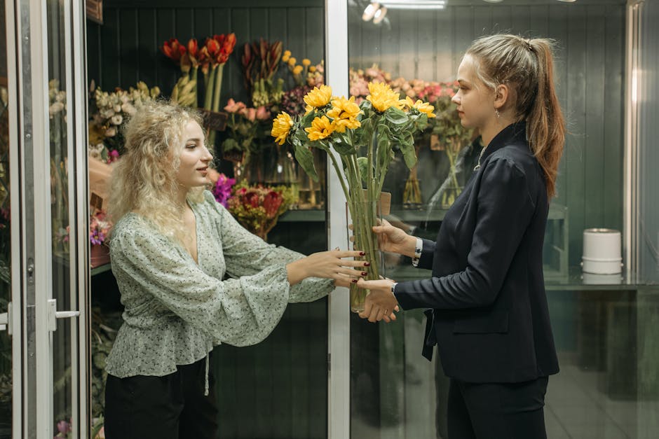 A florist with curly blonde hair, wearing a light green patterned blouse, receives a bouquet of bright yellow sunflowers with green stems from a customer in a floral shop. The customer, with long brow