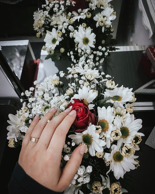 A close-up view of a floral bouquet featuring a single red rose surrounded by white daisies with yellow centers and baby's breath, held gently by a person wearing a dark long-sleeve garment with a sil