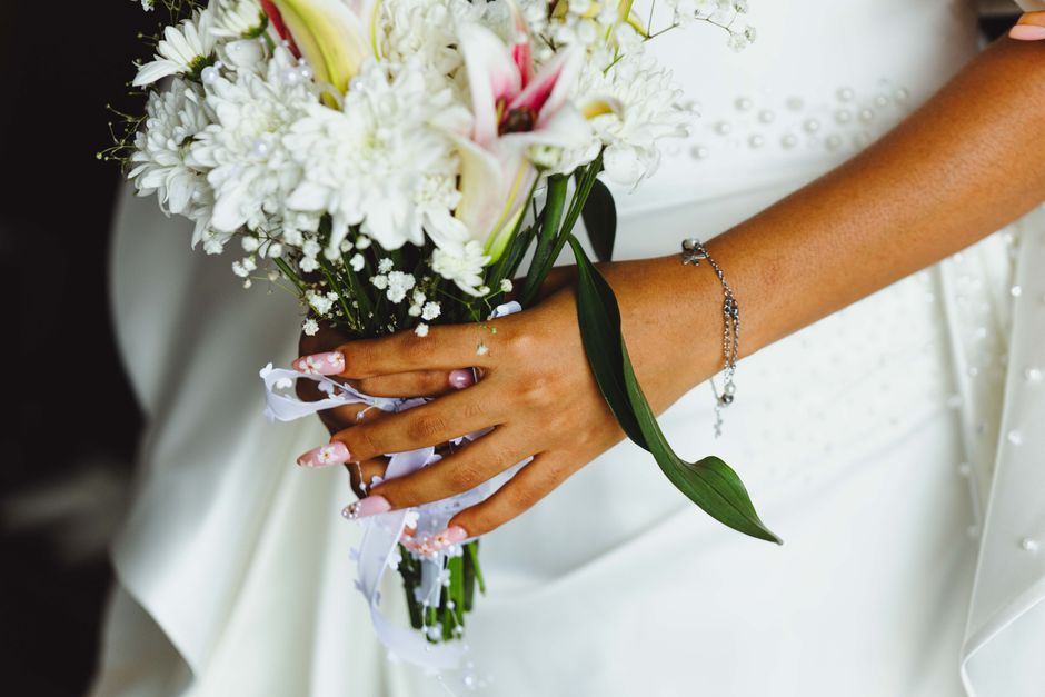 Close-up of a bride’s hand holding a bouquet of fresh white flowers, including lilies, chrysanthemums, and baby's breath, wrapped with white satin ribbons and decorated with a silver bracelet. The b
