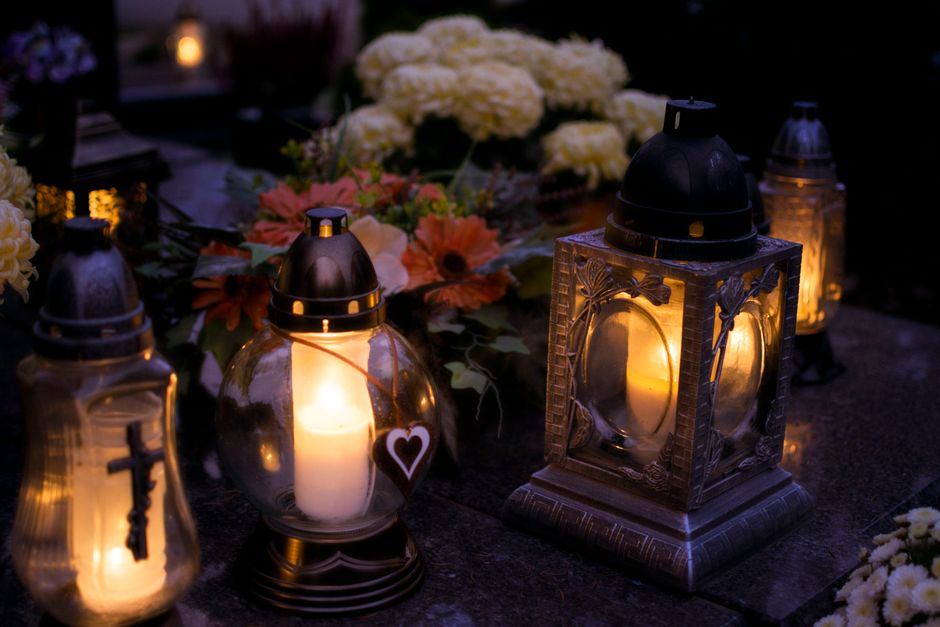A collection of floral arrangements with white and orange flowers, including hydrangeas and gerbera daisies, displayed on a dark surface during dusk. Surrounding the flowers are four lit lanterns with