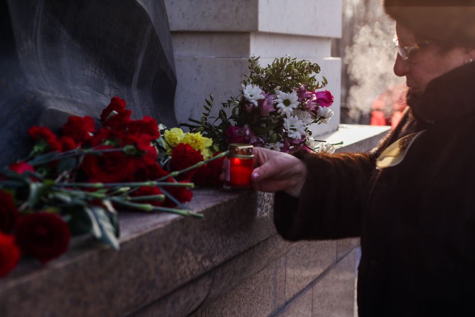 A person placing a lit red candle among an arrangement of fresh flowers for a funeral at a memorial wall. The flowers include red carnations, white daisies, and pink rosebuds, wrapped in clear celloph