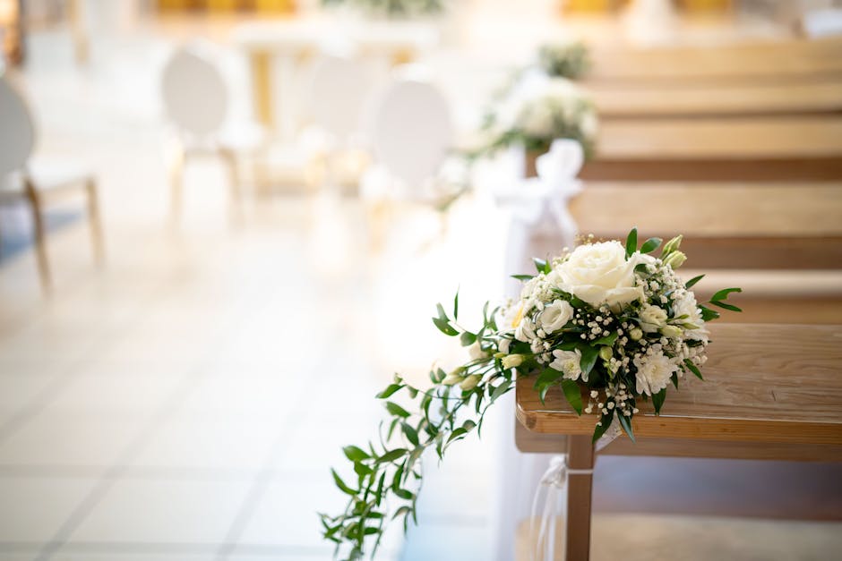 A bouquet of fresh white flowers, including roses and smaller blossoms, decorated with green foliage, resting on a wooden church pew. The arrangement features a cascading design, with some flowers and